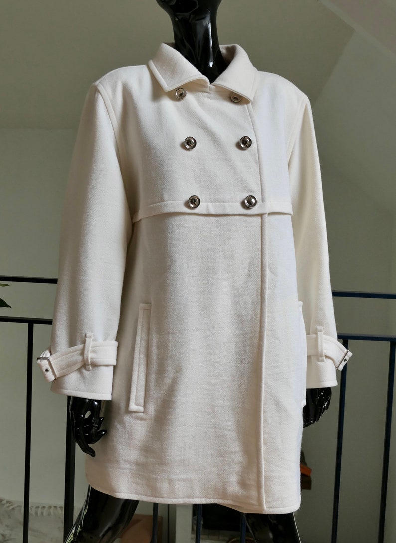 May include: Off-white double-breasted coat with silver-tone buttons. The coat features a notched collar, long sleeves with adjustable cuffs, and two front pockets. The coat is displayed on a black mannequin.