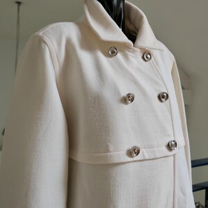 May include: Off-white double-breasted coat with a classic collar and silver-tone buttons. The coat has a textured fabric and a tailored fit. The coat is displayed on a black mannequin.