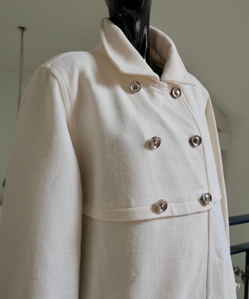 May include: Off-white double-breasted coat with a classic collar and silver-tone buttons. The coat has a textured fabric and a tailored fit. The coat is displayed on a black mannequin.