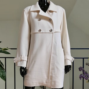 May include: A cream-colored coat with a double-breasted design. The coat features silver buttons, a notched collar, and long sleeves with adjustable cuffs. The coat is displayed on a black mannequin.