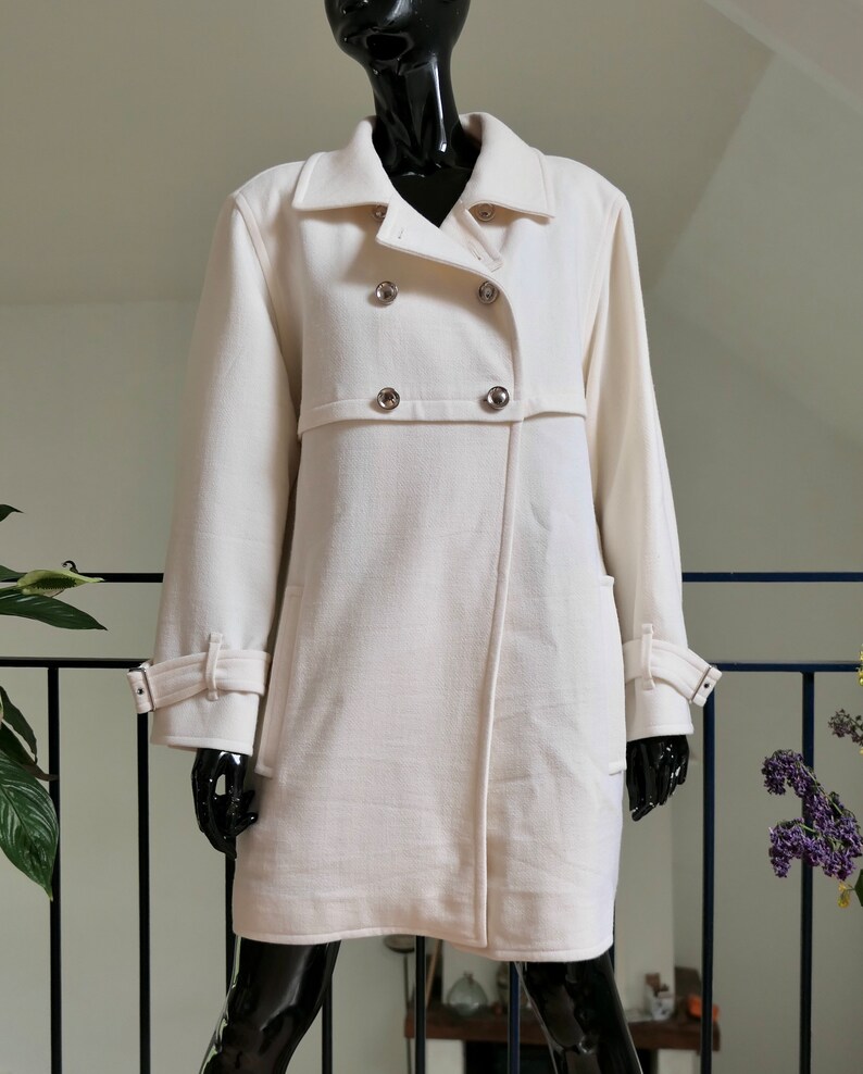 May include: A cream-colored coat with a double-breasted design. The coat features silver buttons, a notched collar, and long sleeves with adjustable cuffs. The coat is displayed on a black mannequin.
