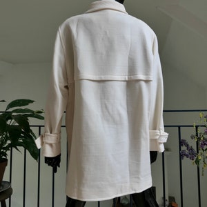 May include: Cream-colored coat with a classic design. The coat features a notched collar, long sleeves with buttoned cuffs, and a back yoke detail. The fabric has a subtle texture, and the coat appears to be a mid-length style.
