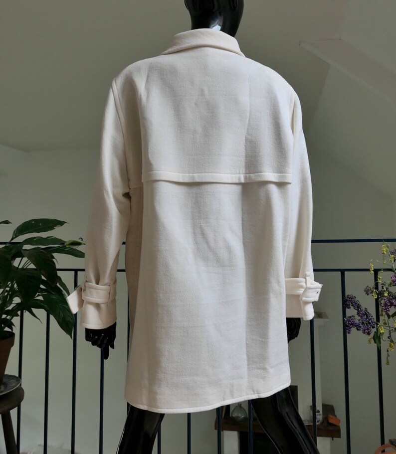 May include: Cream-colored coat with a classic design. The coat features a notched collar, long sleeves with buttoned cuffs, and a back yoke detail. The fabric has a subtle texture, and the coat appears to be a mid-length style.