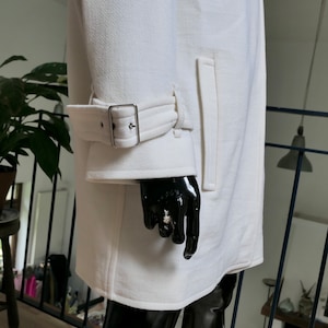 May include: A white coat with a textured surface. The coat features a belt detail on the sleeve with a silver buckle and a vertical pocket. The coat is displayed on a black mannequin.