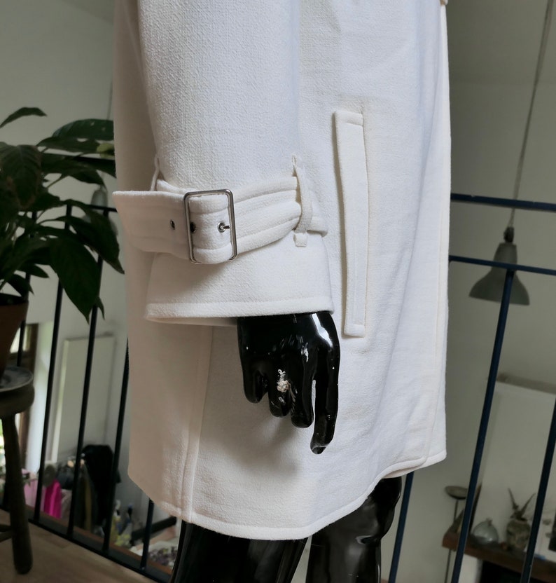 May include: A white coat with a textured surface. The coat features a belt detail on the sleeve with a silver buckle and a vertical pocket. The coat is displayed on a black mannequin.