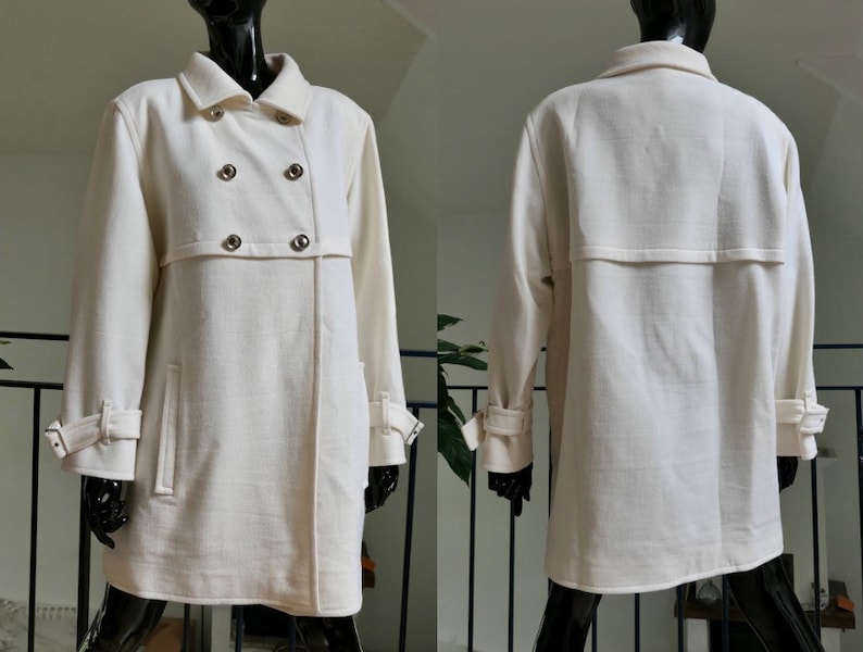 May include: Cream-colored double-breasted coat with silver-tone buttons and a notched collar. The coat features long sleeves with adjustable cuffs and two front pockets. The coat is displayed on a black mannequin.