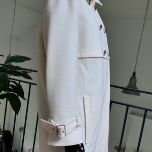 May include: A white coat with a double-breasted design, featuring silver-toned buttons and a notched collar. The coat has a belt detail at the waist and a buckle closure on the sleeves. The coat is displayed on a black mannequin.