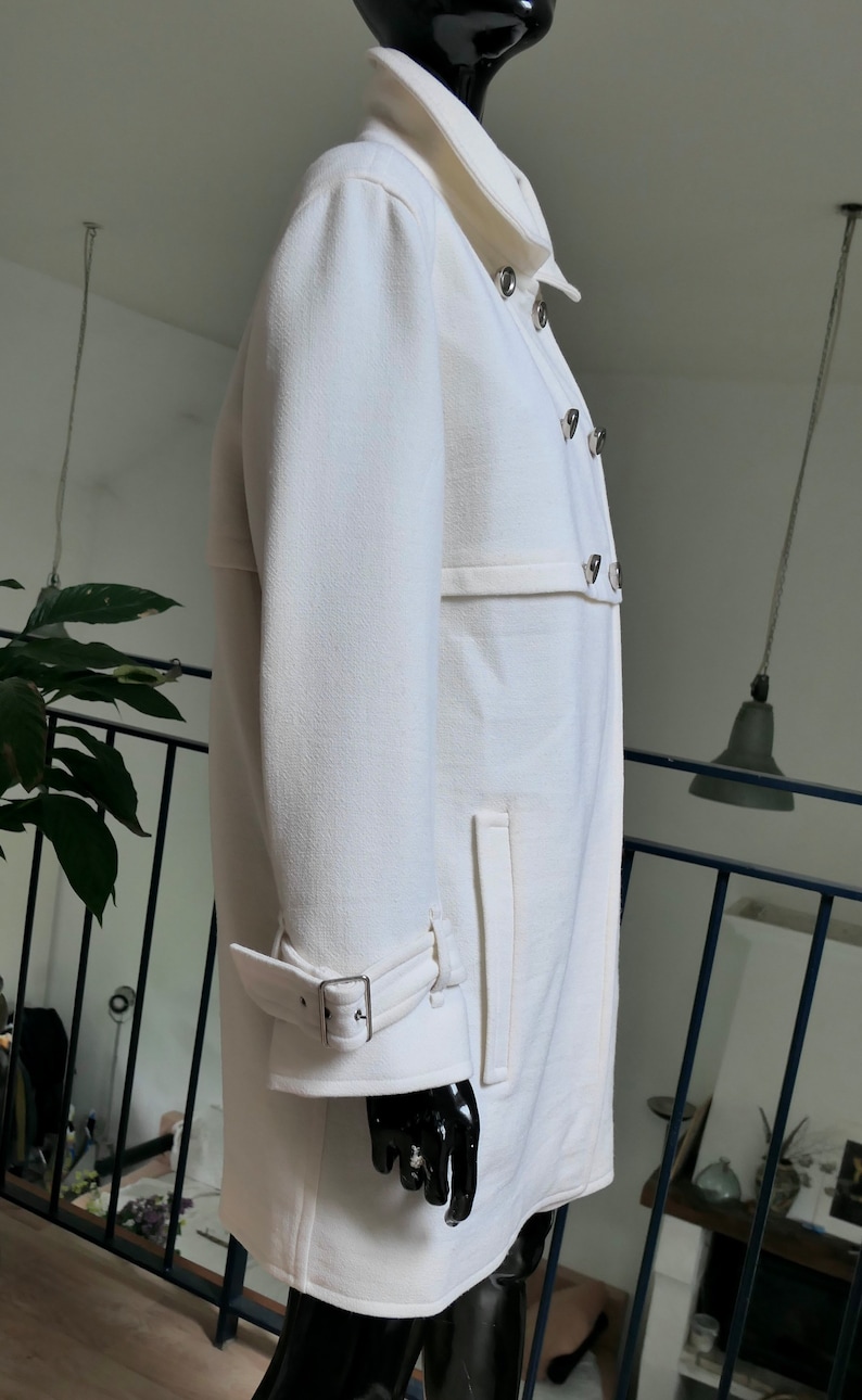 May include: A white coat with a double-breasted design, featuring silver-toned buttons and a notched collar. The coat has a belt detail at the waist and a buckle closure on the sleeves. The coat is displayed on a black mannequin.