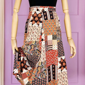 May include: A mannequin wearing a black vest top and a patchwork skirt with a variety of patterns in red, orange, black, and cream. A wooden beaded necklace and a patterned headscarf complete the outfit. The skirt's design is reminiscent of vintage styles.