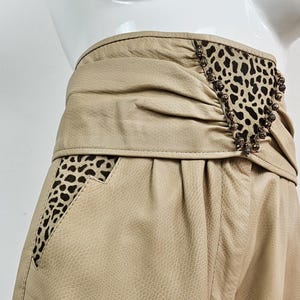 May include: Tan leather trousers with a high waist and distinctive detailing. The waistband incorporates a leopard print section and decorative elements. The trousers have a textured finish and a tailored cut, reflecting a vintage style.