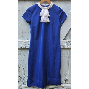 May include: A royal blue dress featuring short sleeves, a white round collar, and a white ruffled jabot detail at the neckline. The fabric has a subtle vertical pinstripe texture, and the garment is displayed on a wooden hanger.
