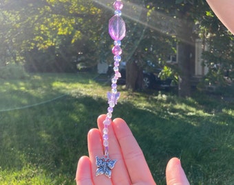 May include: A purple and clear crystal beaded keychain with a silver butterfly charm. The keychain is hanging from a hand.