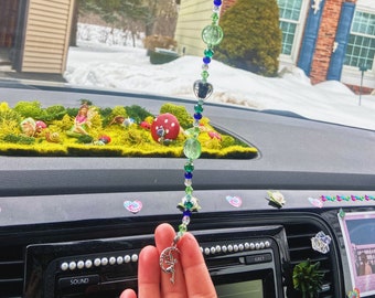May include: A green, blue, and clear crystal beaded car charm with a silver fairy charm hanging from the bottom. The charm is hanging from the rearview mirror of a car.