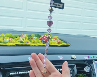 May include: A purple and clear beaded car charm with a gold star charm at the bottom. The charm is hanging from a rearview mirror in a car. The charm has a small tag that says "Black Ice" and a larger tag that says "Little Trees".