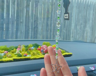 May include: A green and clear beaded car charm with a star charm at the bottom. The charm is hanging from a rearview mirror in a car. The charm also has a small wooden tree charm with the text "Live This" on it.