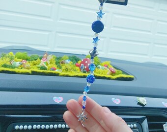 May include: A beaded car charm with blue and clear beads, silver accents, and a snowflake charm. The charm is hanging in front of a car dashboard with a miniature fairy garden.