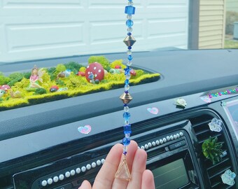May include: A blue and gold beaded car charm with a crystal prism hanging from the bottom. The charm is hanging from a rear view mirror in a car. The charm has a Little Trees air freshener attached to it.