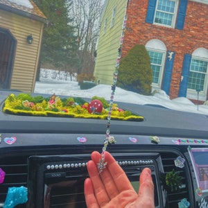 May include: A car interior with a fairy-themed car charm hanging from the rearview mirror. The charm is made of iridescent beads and features a silver fairy figurine. The car dashboard is decorated with a miniature fairy garden and other decorative elements.