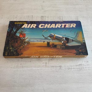 May include: A vintage board game box for "Air Charter" by Waddingtons. The box features a colorful illustration of a plane on a runway with cargo and passengers. The text on the box reads "An absorbing game of air cargo trading. For 2 to 4 players. A Short Game for junior pilots. A Full Game for experienced operators."