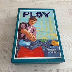 May include: A blue box with the word "PLOY" in white letters. The box features a man in a purple shirt playing a board game with colorful pieces. The text on the box reads "Strategic game of maneuver and capture".