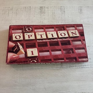 May include: A vintage Parker Brothers Double-Sided Word Game. The red game box displays the word "OPTION" with letter tiles. The game includes a grid for placing tiles and a hand holding a tile. The game is on a light wood surface.