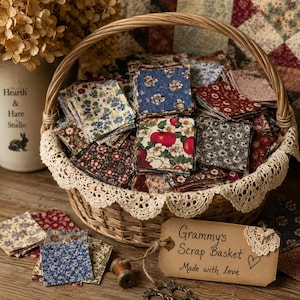 May include: A woven wicker basket filled with assorted square fabric scraps featuring floral and fruit patterns. The basket is lined with a cream-colored lace trim and includes a tan paper tag labeled Grammy's Scrap Basket Made with love.
