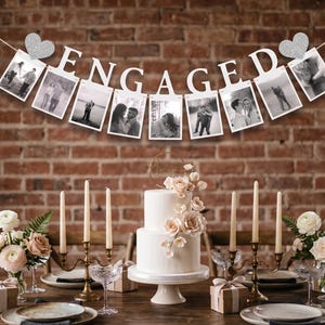 May include: A white two-tier cake decorated with light pink flowers on a stand, flanked by two gold candelabras with lit candles, floral arrangements, and gift boxes. Above, a banner spells ENGAGED with hanging black and white photographs.