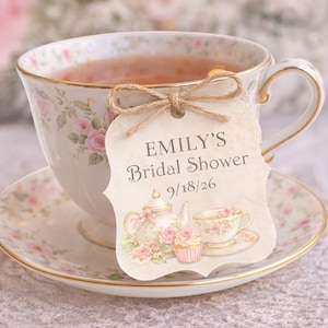May include: A teacup and saucer set with a floral design, filled with tea. A decorative tag with the text "EMILY'S Bridal Shower 9/18/26" is tied to the cup with twine. The set has gold trim.