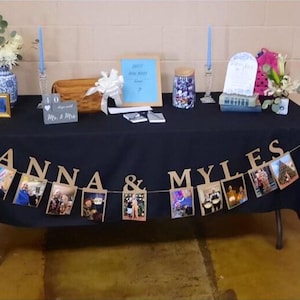 May include: A black table with a black tablecloth and a photo banner that says "Anna & Myles". The banner is decorated with photos of the couple. There are also two candlesticks with blue candles, a small wooden box, a glass jar, and a sign that says "Guess how many kisses?"