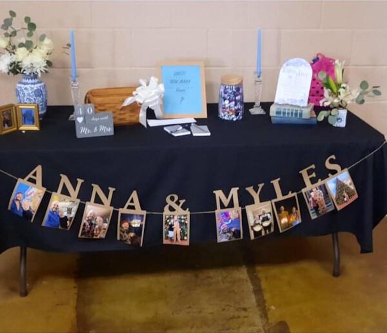May include: A black table with a black tablecloth and a photo banner that says "Anna & Myles". The banner is decorated with photos of the couple. There are also two candlesticks with blue candles, a small wooden box, a glass jar, and a sign that says "Guess how many kisses?"