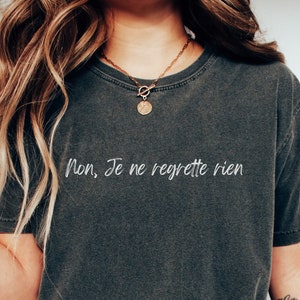 May include: A dark gray t-shirt with the French phrase "Non, Je ne regrette rien" printed in white on the front. The shirt is worn by a person with long blonde hair.