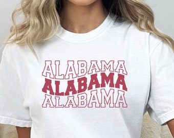 May include: White t-shirt with the word "ALABAMA" repeated three times in a red, outlined font. The text is stacked vertically, with each word slightly offset from the one above it.