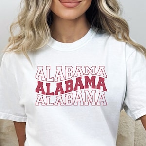 May include: White t-shirt with the word "ALABAMA" repeated three times in a red, outlined font. The text is stacked vertically, with each word slightly offset from the one above it.