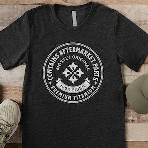 Puede incluir: Una camiseta gris oscuro con un gr&aacute;fico circular blanco que dice "Contiene piezas de recambio - Mayormente originales - 100% Bi&oacute;nico - Titanio de primera calidad".