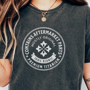 Peut inclure: Un t-shirt gris foncé avec un imprimé graphique blanc en forme de cercle avec le texte "Contains Aftermarket Parts" et "Mostly Original" et "100% Bionic" et "Premium Titanium".