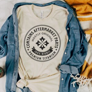 Puede incluir: Una camiseta beige con un gr&aacute;fico circular negro que dice "Contiene piezas de recambio" con las palabras "Mayormente original" y "100% Bi&oacute;nico Titanio Premium" alrededor del c&iacute;rculo.