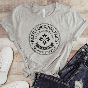 Peut inclure: T-shirt gris avec un graphique circulaire noir qui dit "Mostly Original Parts" avec les mots "Aftermarket Hip" et "100% Bionic Premium Titanium" autour du cercle.