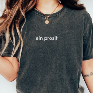 May include: A dark gray cropped t-shirt with the text "ein prosit" printed on the front. The shirt is worn by a person with long brown hair and a gold necklace with a circular pendant.