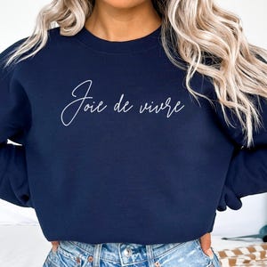 May include: Navy blue sweatshirt with the phrase "Joie de vivre" in white script. The long-sleeved crew neck top is made of a soft material. The sweatshirt is a casual style.