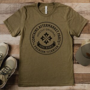 Puede incluir: Una camiseta verde con un gr&aacute;fico circular negro que dice "Contiene piezas de recambio" con un dise&ntilde;o de flor estilizado y el texto "Mayormente original, 100% Bi&oacute;nico, Titanio de primera calidad".
