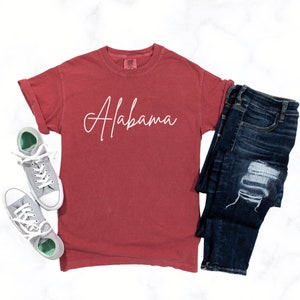 May include: A red t-shirt with the word "Alabama" written in white cursive on the front. The shirt is paired with blue jeans and gray sneakers.
