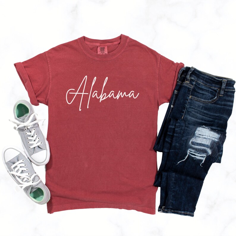 May include: A red t-shirt with the word "Alabama" written in white cursive on the front. The shirt is paired with blue jeans and gray sneakers.