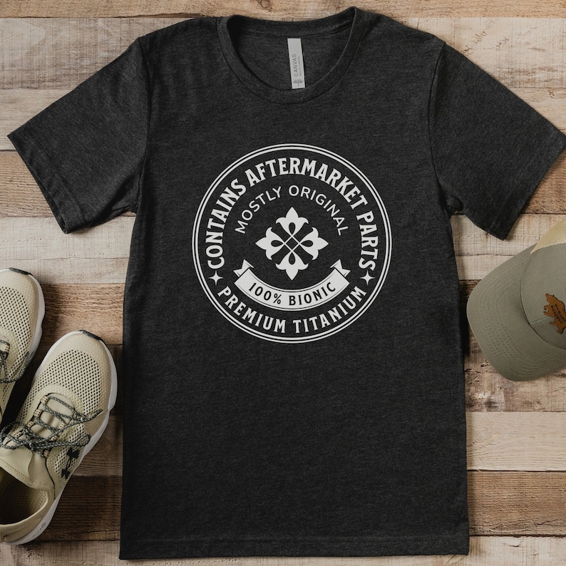 Puede incluir: Una camiseta gris oscuro con un gr&aacute;fico circular blanco que dice "Contiene piezas de recambio - Mayormente originales - 100% Bi&oacute;nico - Titanio de primera calidad".