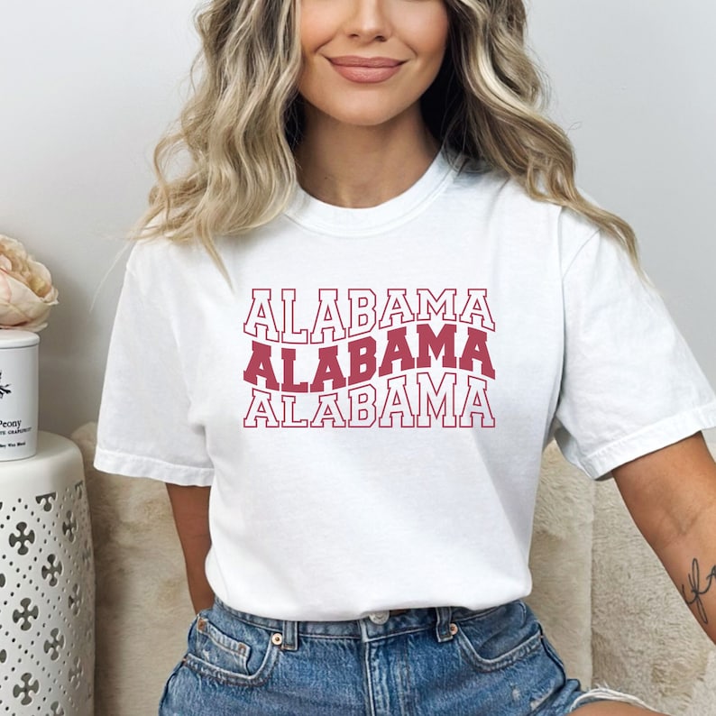 May include: White t-shirt with the word "ALABAMA" repeated three times in a red, outlined font. The text is stacked vertically, with each word slightly offset from the one above it.