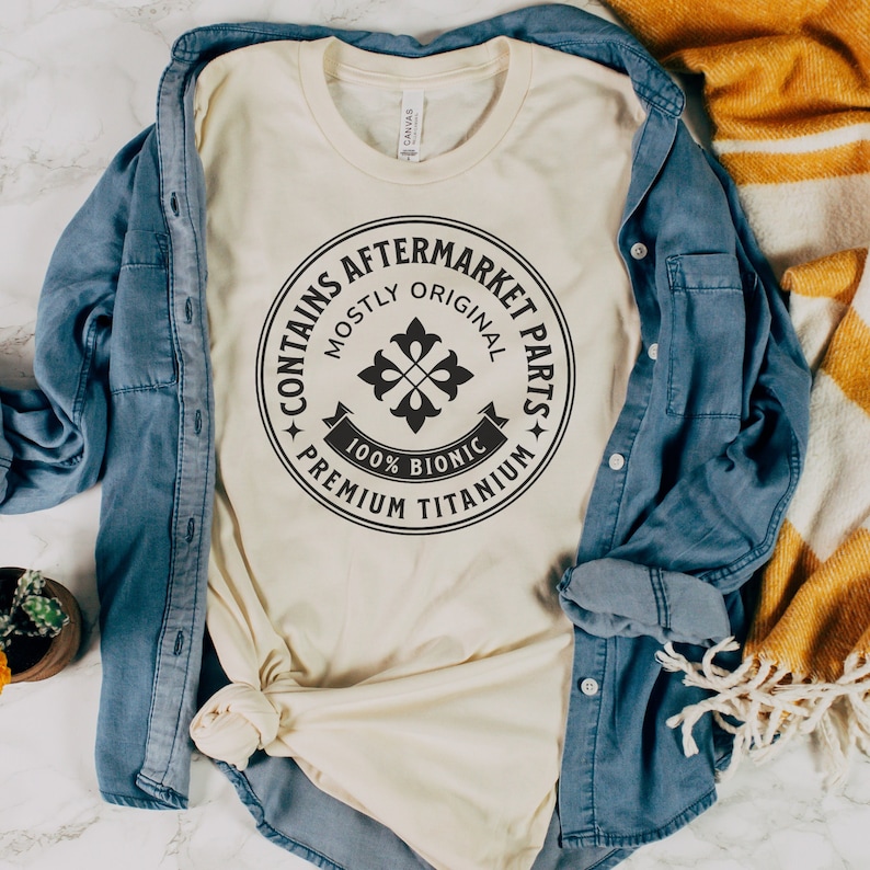 Puede incluir: Una camiseta beige con un gr&aacute;fico circular negro que dice "Contiene piezas de recambio" con las palabras "Mayormente original" y "100% Bi&oacute;nico Titanio Premium" alrededor del c&iacute;rculo.