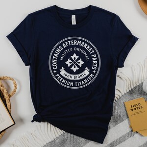 Puede incluir: Una camiseta azul marino con un gr&aacute;fico circular blanco que dice "Contiene piezas de recambio" con el texto "Mayormente original" y "100% Bi&oacute;nico" dentro del c&iacute;rculo. El c&iacute;rculo tambi&eacute;n tiene el texto "Titanio de primera calidad" alrededor del exterior.