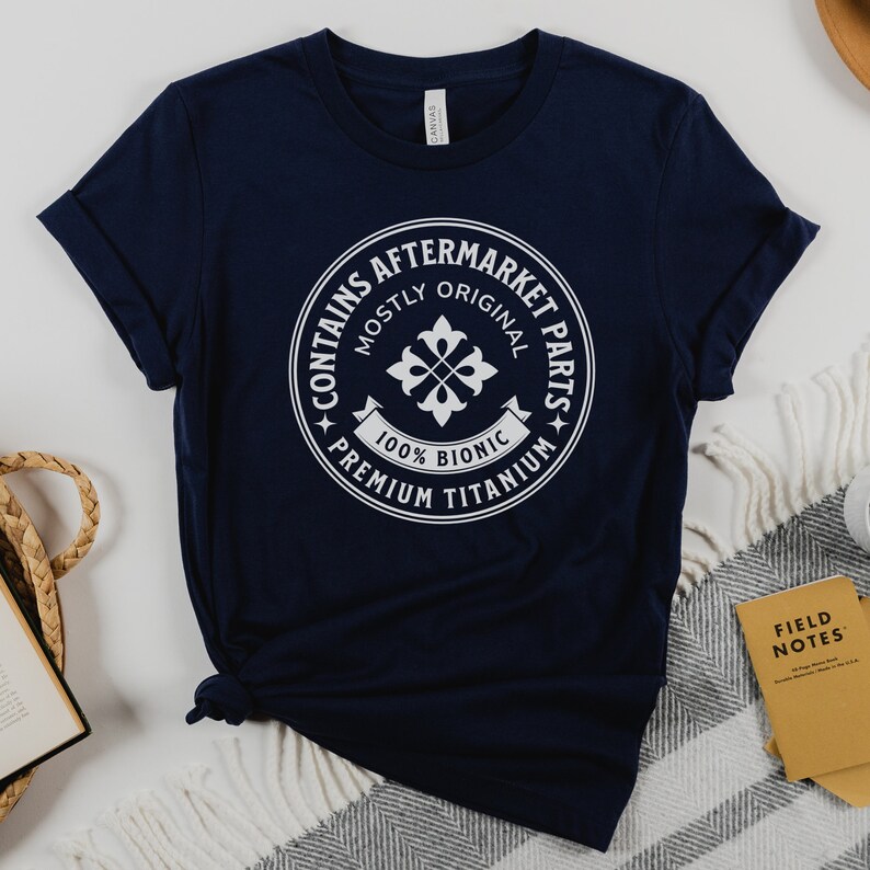 Puede incluir: Una camiseta azul marino con un gr&aacute;fico circular blanco que dice "Contiene piezas de recambio" con el texto "Mayormente original" y "100% Bi&oacute;nico" dentro del c&iacute;rculo. El c&iacute;rculo tambi&eacute;n tiene el texto "Titanio de primera calidad" alrededor del exterior.