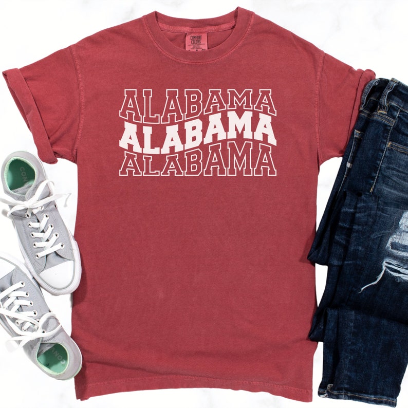 May include: A red t-shirt with the word "ALABAMA" printed three times in white, stacked vertically. The shirt is folded and laid flat on a white surface. A pair of blue jeans is folded and laid flat to the right of the shirt.