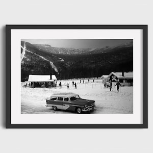 May include: A black and white photo of a vintage station wagon parked in a snowy mountain setting.  People are skiing and walking in the background.  There are two small wooden buildings in the background.