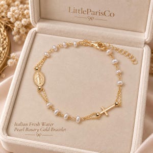May include: A gold rosary bracelet featuring small white freshwater pearls, a central oval medal depicting a figure, and a small gold cross charm. The bracelet includes a delicate gold chain with a lobster clasp and an adjustable extender link chain.